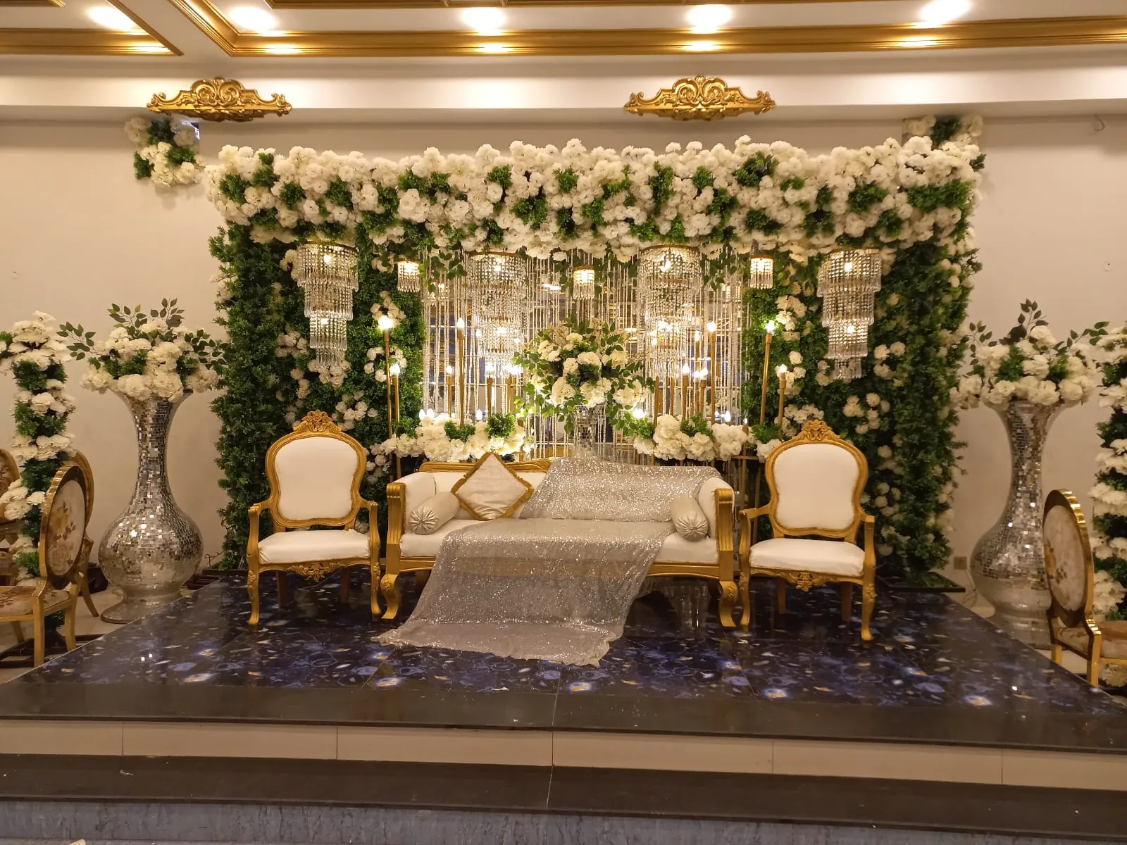 NURI bridal stage with a white rose wall, crystal chandeliers and a gold-framed white sofa